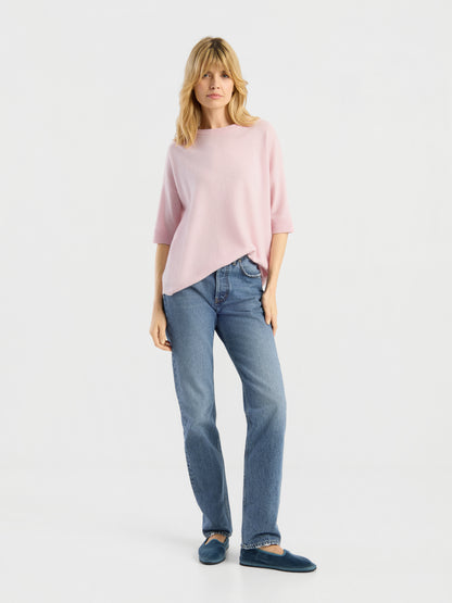 Vor einem schlichten weißen Hintergrund steht eine Frau mit blonden Haaren und einem entspannten Gesichtsausdruck. Sie trägt das Strick-T-Shirt „Aurora Rose“, eine blaue Jeans und dunkelblaue Hausschuhe.