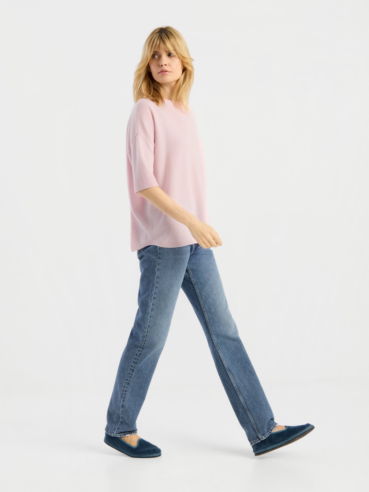 Eine Frau mit blonden Haaren trägt das Strick-T-Shirt „Aurora Rose“, dazu blaue Jeans und blaue Schuhe, und geht vor einem weißen Hintergrund nach links.