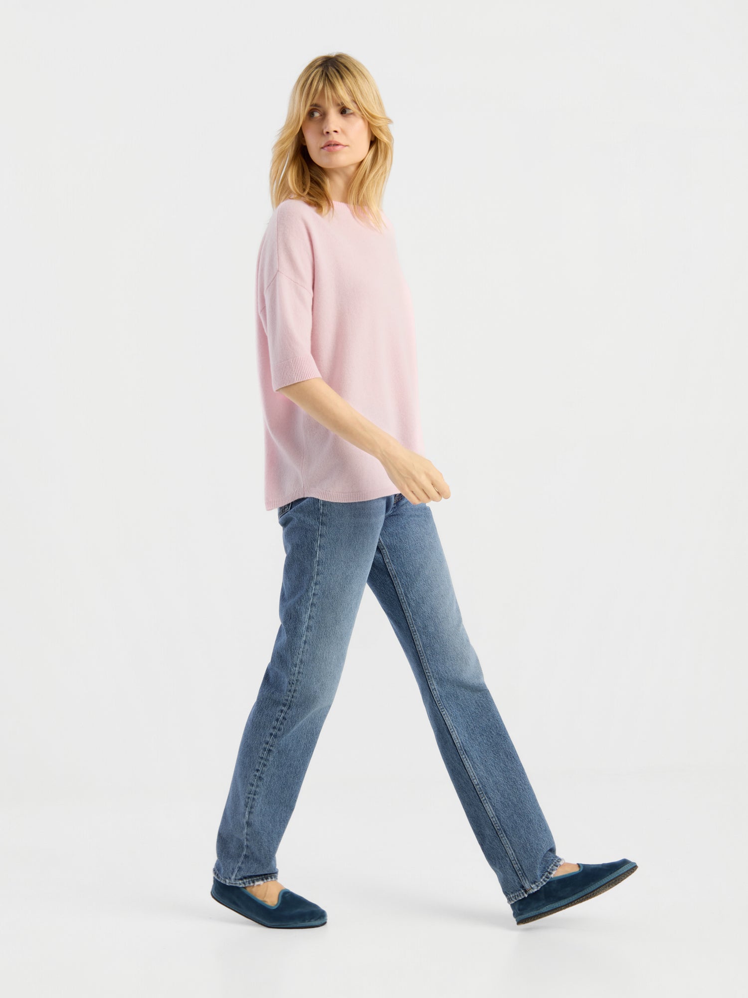 Eine Frau mit blonden Haaren trägt das Strick-T-Shirt „Aurora Rose“, dazu blaue Jeans und blaue Schuhe, und geht vor einem weißen Hintergrund nach links.