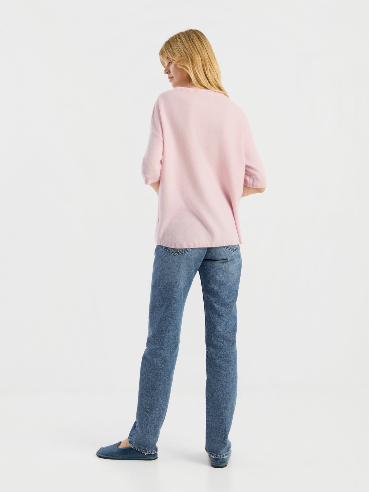 Eine Person steht mit dem Rücken zur Kamera und trägt das Strick-T-Shirt „Aurora Rose“, eine blaue Jeans und blaue Schuhe vor einem schlichten weißen Hintergrund.