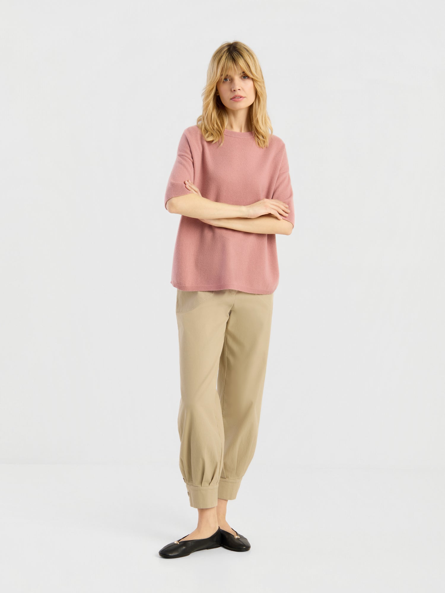 Eine Person steht vor einem schlichten weißen Hintergrund und trägt das Strick-T-Shirt „Aurora Altrosa“ sowie eine hellbraune Hose mit Umschlag, dazu schwarze Ballerinas. Sie hat blondes Haar und einen neutralen Gesichtsausdruck.