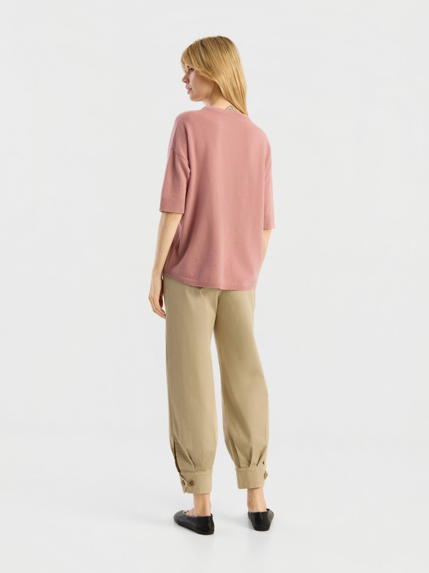 Vor einem schlichten weißen Hintergrund steht eine Frau mit dem Rücken zur Kamera. Sie trägt ein lockeres, kurzärmeliges Strick-T-Shirt der Marke „Aurora Altrosa“ in Rosa, dazu eine beige Hose mit Umschlag und schwarze Schuhe.