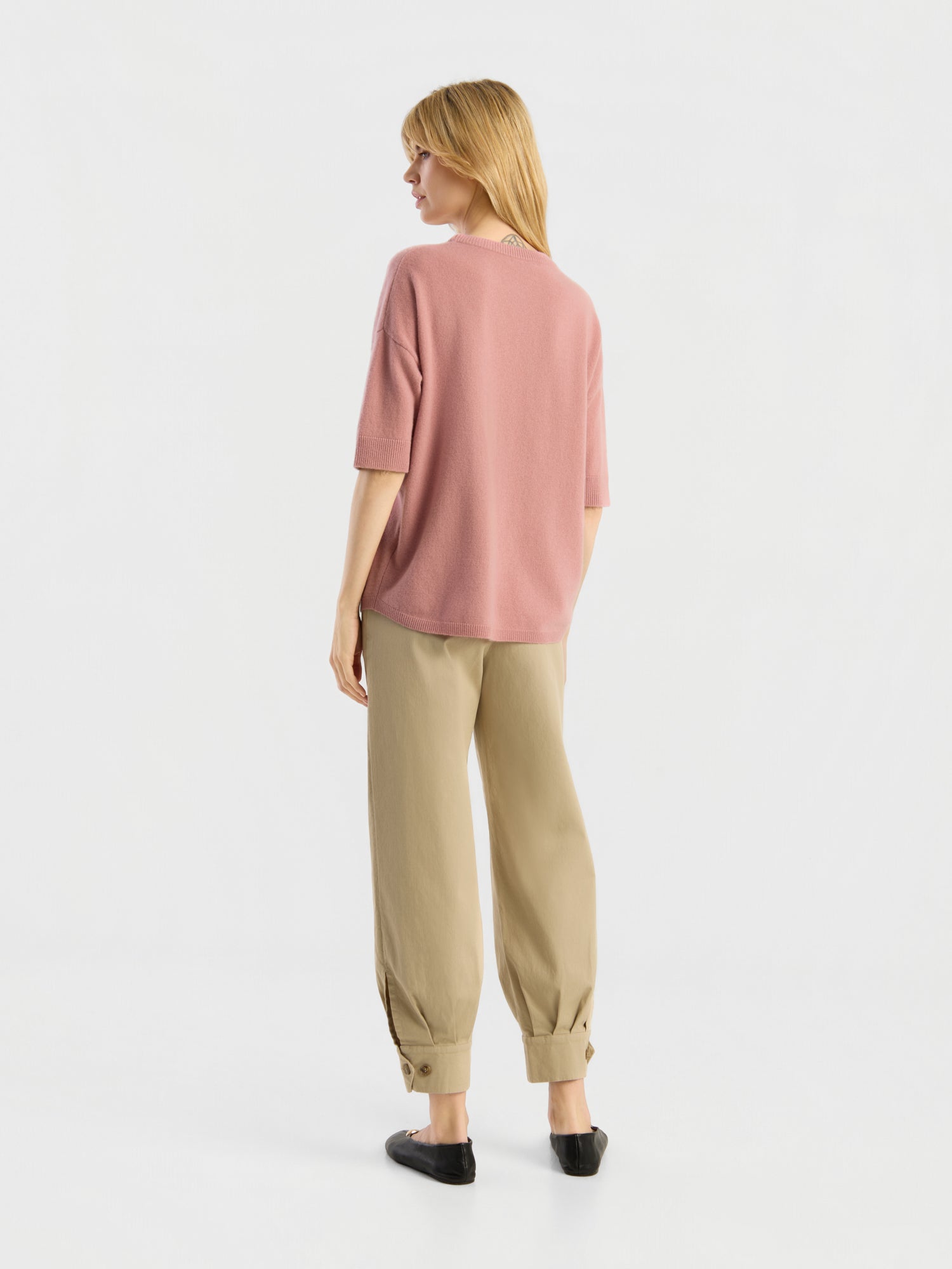 Vor einem schlichten weißen Hintergrund steht eine Frau mit dem Rücken zur Kamera. Sie trägt ein lockeres, kurzärmeliges Strick-T-Shirt der Marke „Aurora Altrosa“ in Rosa, dazu eine beige Hose mit Umschlag und schwarze Schuhe.