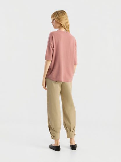 Vor einem schlichten weißen Hintergrund steht eine Frau mit dem Rücken zur Kamera. Sie trägt ein lockeres, kurzärmeliges Strick-T-Shirt der Marke „Aurora Altrosa“ in Rosa, dazu eine beige Hose mit Umschlag und schwarze Schuhe.