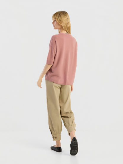 Vor einem schlichten weißen Hintergrund steht eine Person mit dem Rücken zur Kamera und trägt ein locker sitzendes Strick-T-Shirt von Aurora Altrosa, eine beige Hose mit Umschlag und schwarze Schuhe.