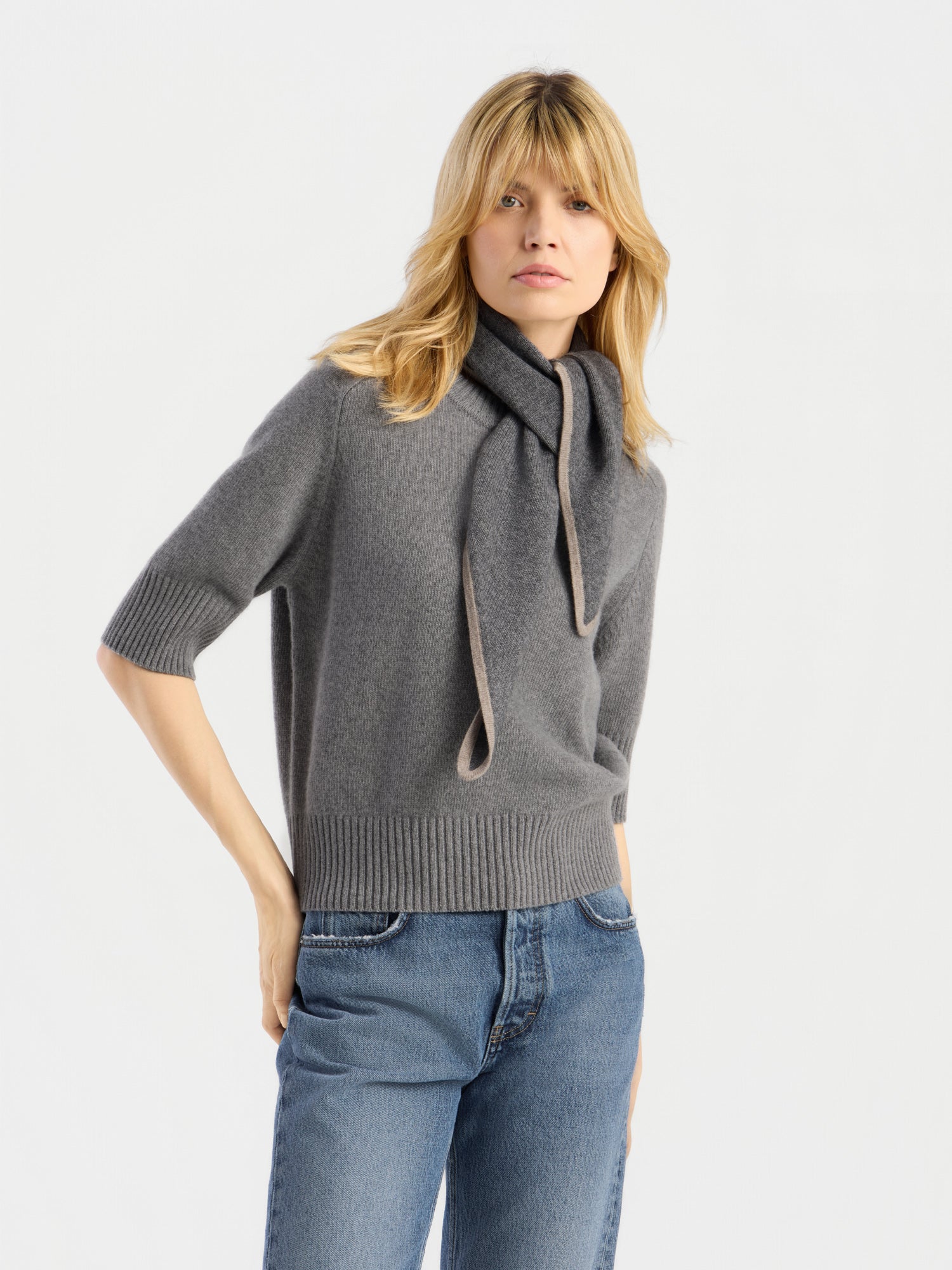 Eine Frau mit blonden Haaren trägt das graue, kurzärmelige Joana-T-Shirt mit einem gebundenen Schal-Kragen und dazu eine blaue Jeans. Vor einem schlichten, hellen Hintergrund posiert sie mit einer Hand in der Tasche.