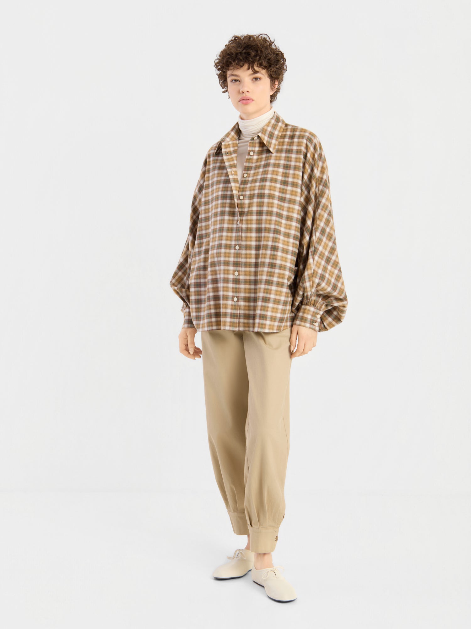 Eine Person, die eine locker sitzende braune Bluse „Antonia Flanell Check“ über einem weißen Rollkragenpullover, einer beigen Jogginghose und weißen Slippern trägt, steht vor einem schlichten weißen Hintergrund.