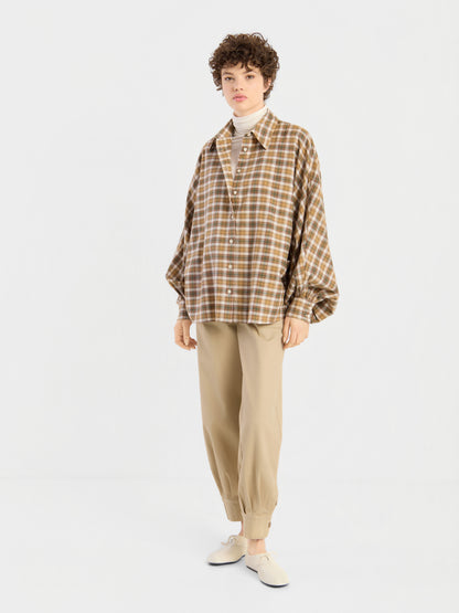 Eine Person, die eine locker sitzende braune Bluse „Antonia Flanell Check“ über einem weißen Rollkragenpullover, einer beigen Jogginghose und weißen Slippern trägt, steht vor einem schlichten weißen Hintergrund.