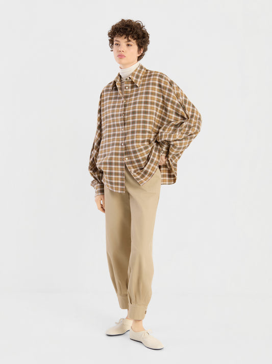 Diese Person steht vor einem schlichten weißen Hintergrund und trägt das braune Karohemd „Falling for Flannel“ über einem cremefarbenen Rollkragenpullover, dazu eine beige Hose mit Umschlag am Knöchel und cremefarbene Slipper.