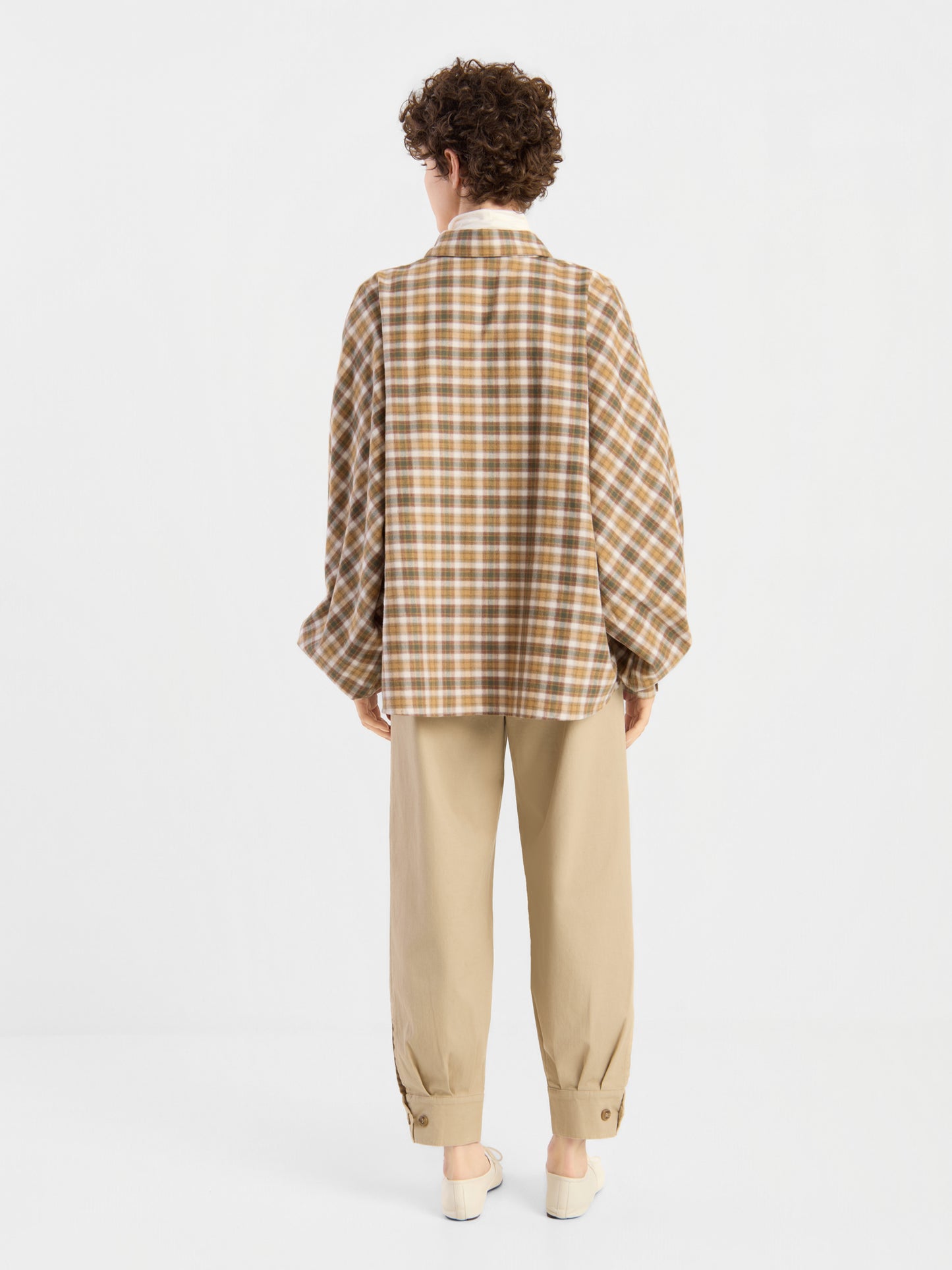 Eine Person mit kurzen Locken, die eine braune Bluse „Antonia Flanell Check“, eine beige Hose und weiße Schuhe trägt, steht mit dem Rücken zur Kamera vor einem schlichten weißen Hintergrund.