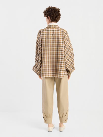 Eine Person mit kurzen Locken, die eine braune Bluse „Antonia Flanell Check“, eine beige Hose und weiße Schuhe trägt, steht mit dem Rücken zur Kamera vor einem schlichten weißen Hintergrund.