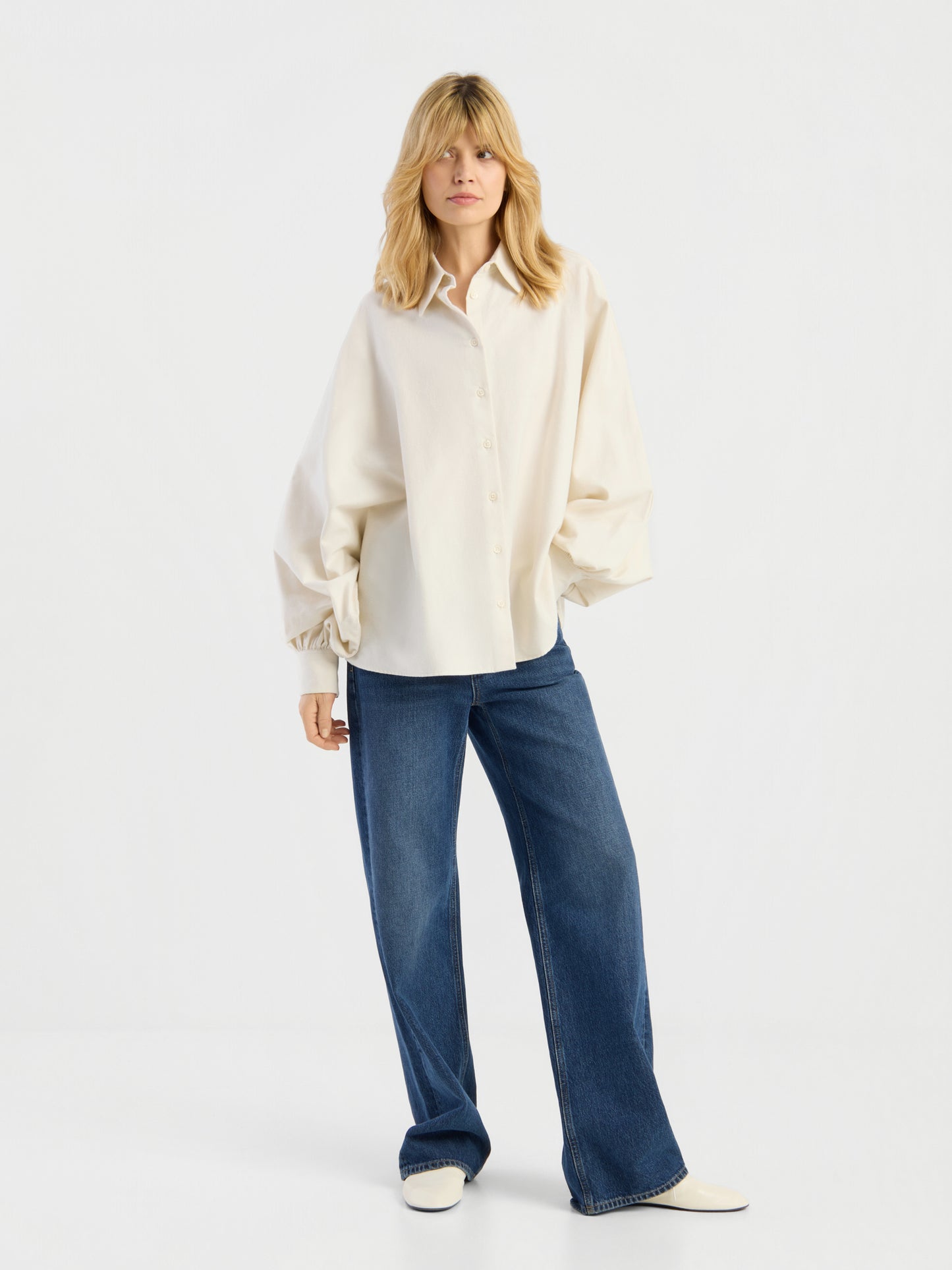 Eine Person, die die Bluse „Antonia Flanell Offwhite“, eine blaue Jeans mit weitem Bein und weiße Schuhe trägt, steht vor einem schlichten weißen Hintergrund.