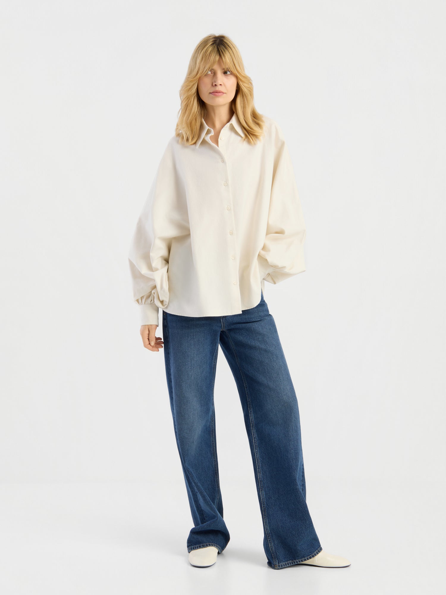Eine Person, die die Bluse „Antonia Flanell Offwhite“, eine blaue Jeans mit weitem Bein und weiße Schuhe trägt, steht vor einem schlichten weißen Hintergrund.