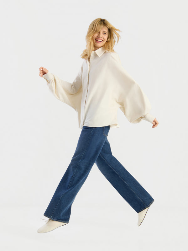 Eine Person mit blonden Haaren, die die Bluse „Blouse in Motion“, eine blaue Jeans mit weitem Bein und weiße Schuhe trägt, lächelt und geht voller Elan vor einem schlichten weißen Hintergrund.