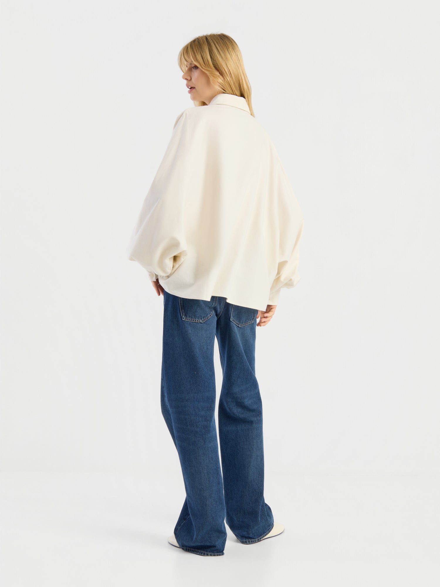Eine Person mit blonden Haaren, die eine locker sitzende, cremefarbene Bluse „Antonia Flanell Offwhite“ und eine blaue Jeans trägt, steht mit dem Rücken zur Kamera vor einem schlichten weißen Hintergrund.