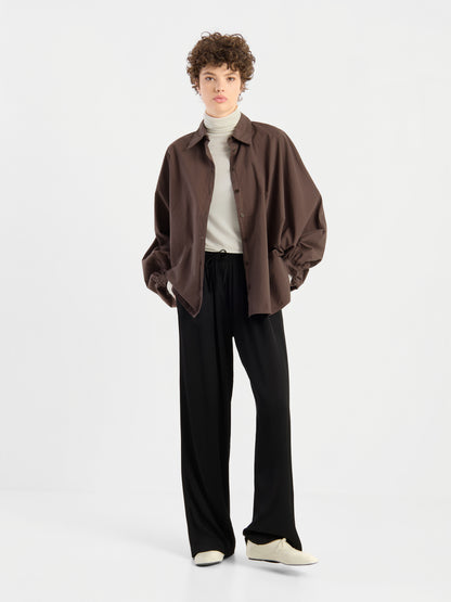 Eine Person mit kurzen Locken trägt einen weißen Rollkragenpullover, die Bluse „Antonia Cotton Espresso“ (Einheitsgröße) als locker sitzendes braunes Hemd mit Knöpfen, eine schwarze Hose mit weitem Bein und weiße Schuhe und steht vor einem schlichten weißen Hintergrund.