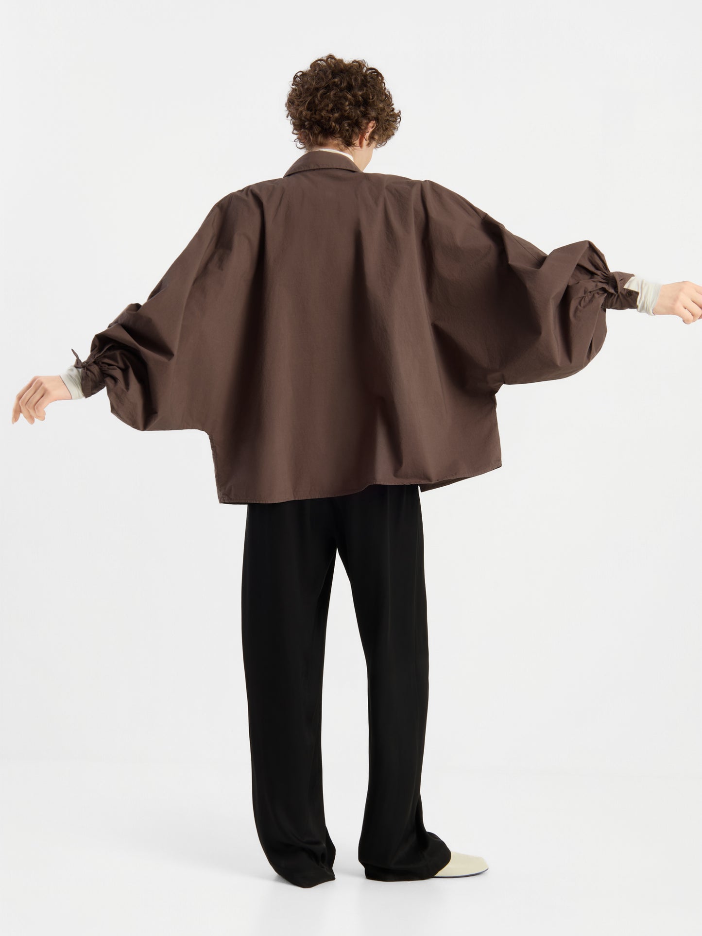 Eine Person steht mit ausgestreckten Armen vor einem schlichten weißen Hintergrund, den Rücken zur Kamera gewandt, und trägt das Modell „Bluse Antonia Cotton Espresso One Size“ – ein übergroßes braunes Hemd mit Puffärmeln –, dazu eine schwarze Hose.