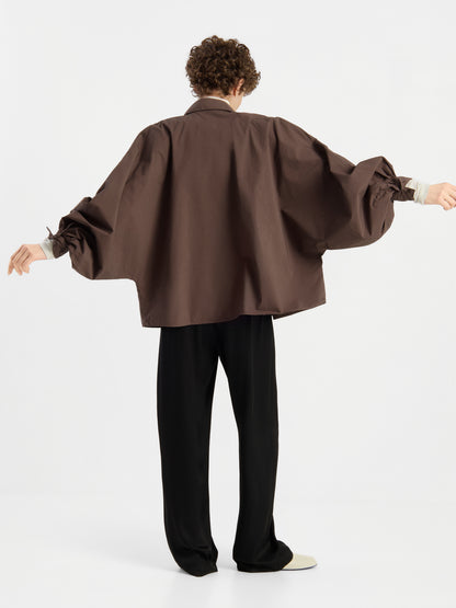Eine Person steht mit ausgestreckten Armen vor einem schlichten weißen Hintergrund, den Rücken zur Kamera gewandt, und trägt das Modell „Bluse Antonia Cotton Espresso One Size“ – ein übergroßes braunes Hemd mit Puffärmeln –, dazu eine schwarze Hose.