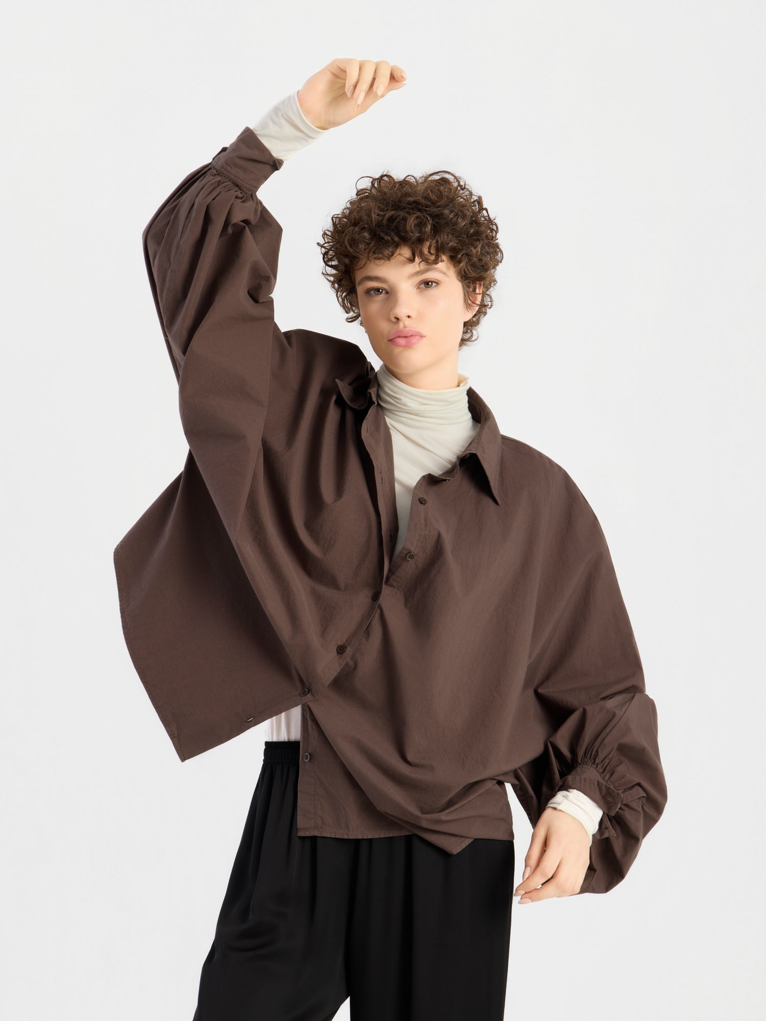 Eine Person mit kurzen Locken trägt die Bluse „Antonia Cotton Espresso“ in Einheitsgröße über einem hellen Rollkragenpullover und einer schwarzen Hose und steht vor einem schlichten weißen Hintergrund, wobei sie einen Arm über den Kopf hebt.