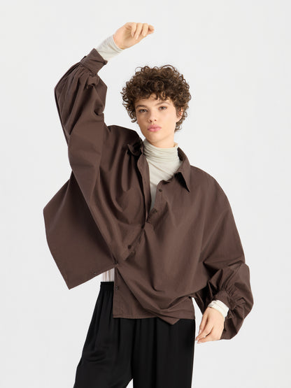 Eine Person mit kurzen Locken trägt die Bluse „Antonia Cotton Espresso“ in Einheitsgröße über einem hellen Rollkragenpullover und einer schwarzen Hose und steht vor einem schlichten weißen Hintergrund, wobei sie einen Arm über den Kopf hebt.