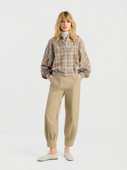 Eine Frau blickt nach vorne und trägt die Bluse „Antonia Short Flanell Check“ über einem hellen Rollkragenpullover, dazu eine lockere beige Hose mit Umschlag und cremefarbene Schuhe; das Ganze vor einem schlichten weißen Hintergrund.