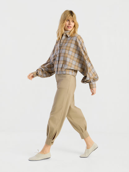 Eine blonde Frau trägt die Bluse „Antonia Short Flanell Check“ mit weiten Ärmeln, eine beige Hose mit hoher Taille und geknöpften Bündchen sowie weiße flache Schuhe und schreitet selbstbewusst vor einem schlichten weißen Hintergrund voran.