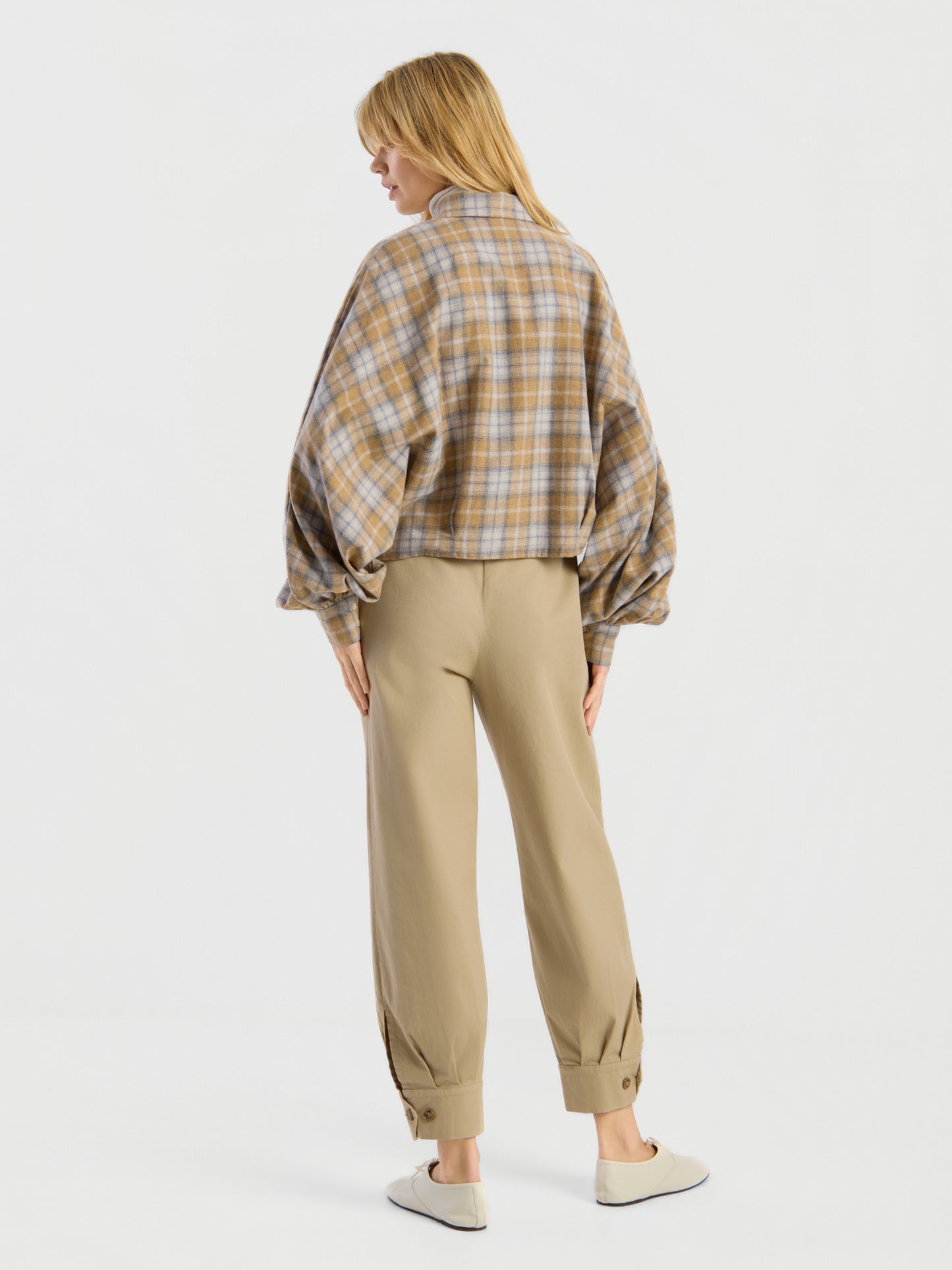 Vor einem schlichten, hellen Hintergrund steht eine Person mit dem Rücken zur Kamera. Sie trägt die Bluse „Antonia Short Flanell Check“ mit Ballonärmeln, eine beige Hose mit Umschlag und Knöpfen am Knöchel sowie weiße Slipper.