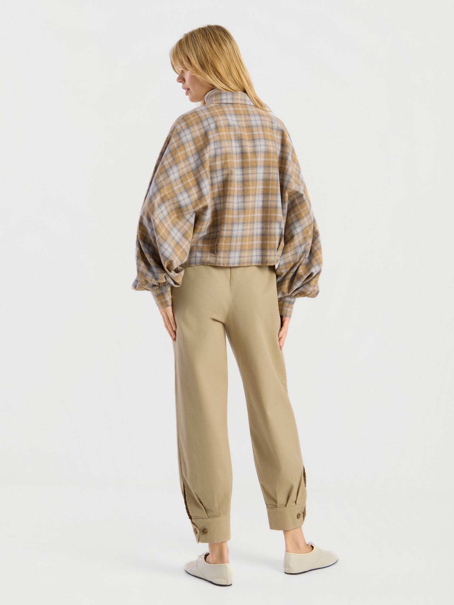 Vor einem schlichten, hellen Hintergrund steht eine Person mit dem Rücken zur Kamera. Sie trägt die Bluse „Antonia Short Flanell Check“ mit Ballonärmeln, eine beige Hose mit Umschlag und Knöpfen am Knöchel sowie weiße Slipper.
