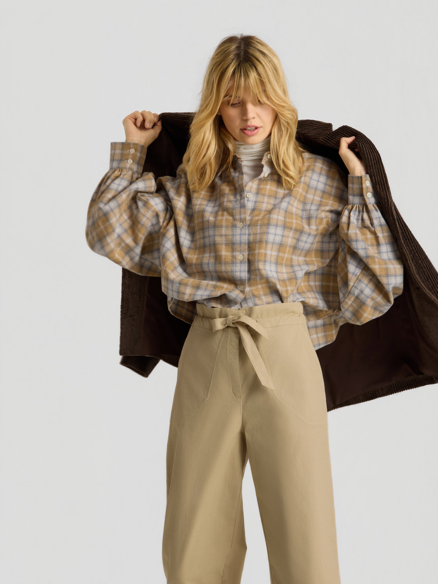 Vor einem schlichten Hintergrund trägt eine blonde Frau die Bluse „Antonia Short Flanell Check“, eine beige Hose mit hoher Taille und gebundenem Gürtel sowie einen dunkelbraunen Mantel, der über ihre Schultern drapiert ist.