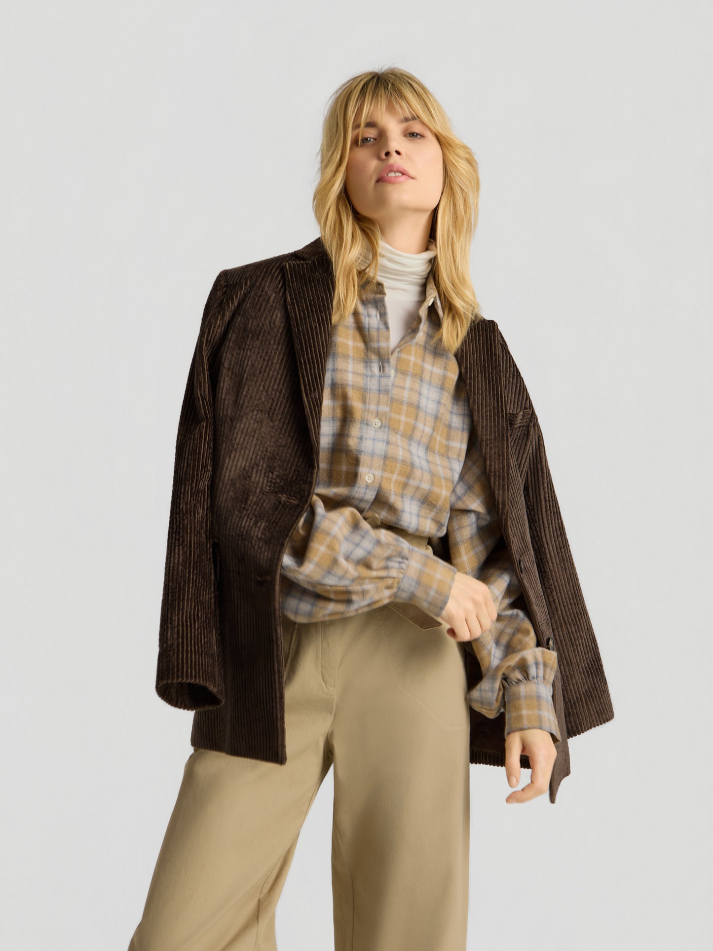 Eine blonde Frau posiert selbstbewusst vor einem schlichten hellgrauen Hintergrund. Sie trägt einen braunen Cordblazer über den Schultern, die Bluse „Antonia Short Flanell Check“ über einem weißen Rollkragenpullover sowie eine beige Hose.