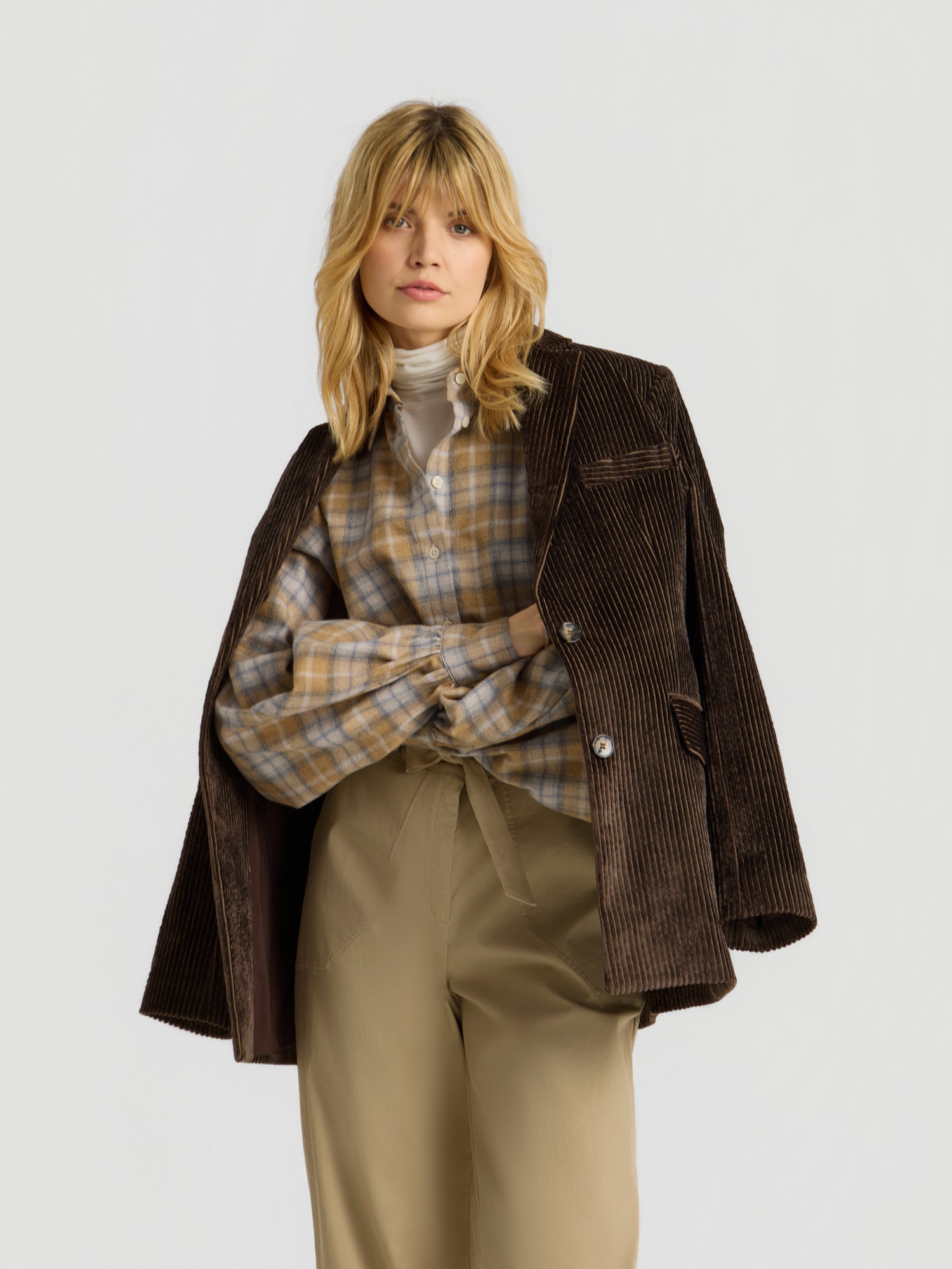 Eine Frau mit blonden Haaren trägt einen braunen Cordblazer über den Schultern, die Bluse „Antonia Short Flanell Check“, einen weißen Rollkragenpullover und eine beige Hose mit hoher Taille, während sie vor einem schlichten, hellen Hintergrund posiert.