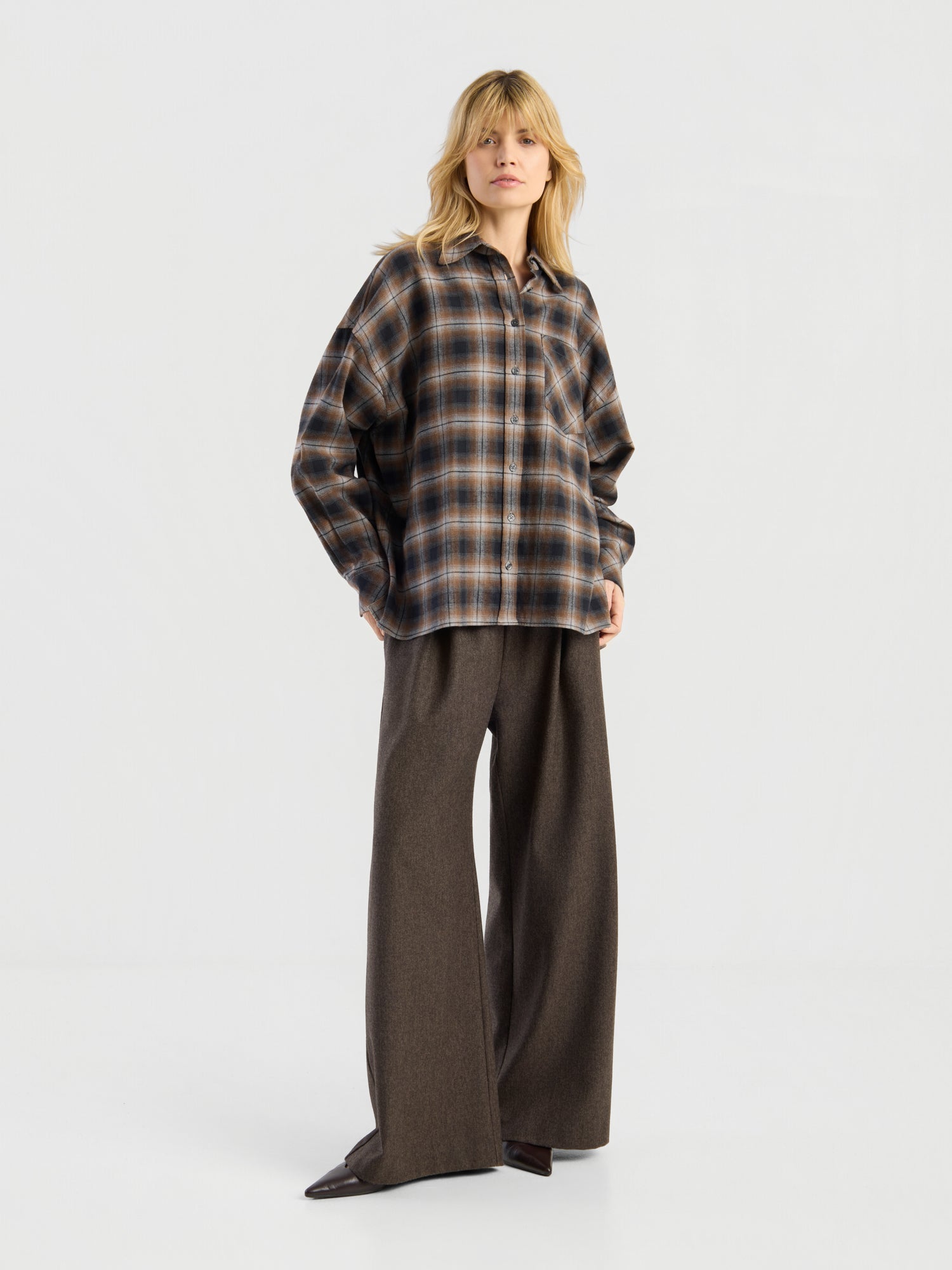 Eine blonde Person trägt die Bluse „Boxy Long“ und eine braune Hose mit weitem Bein und steht vor einem schlichten weißen Hintergrund.
