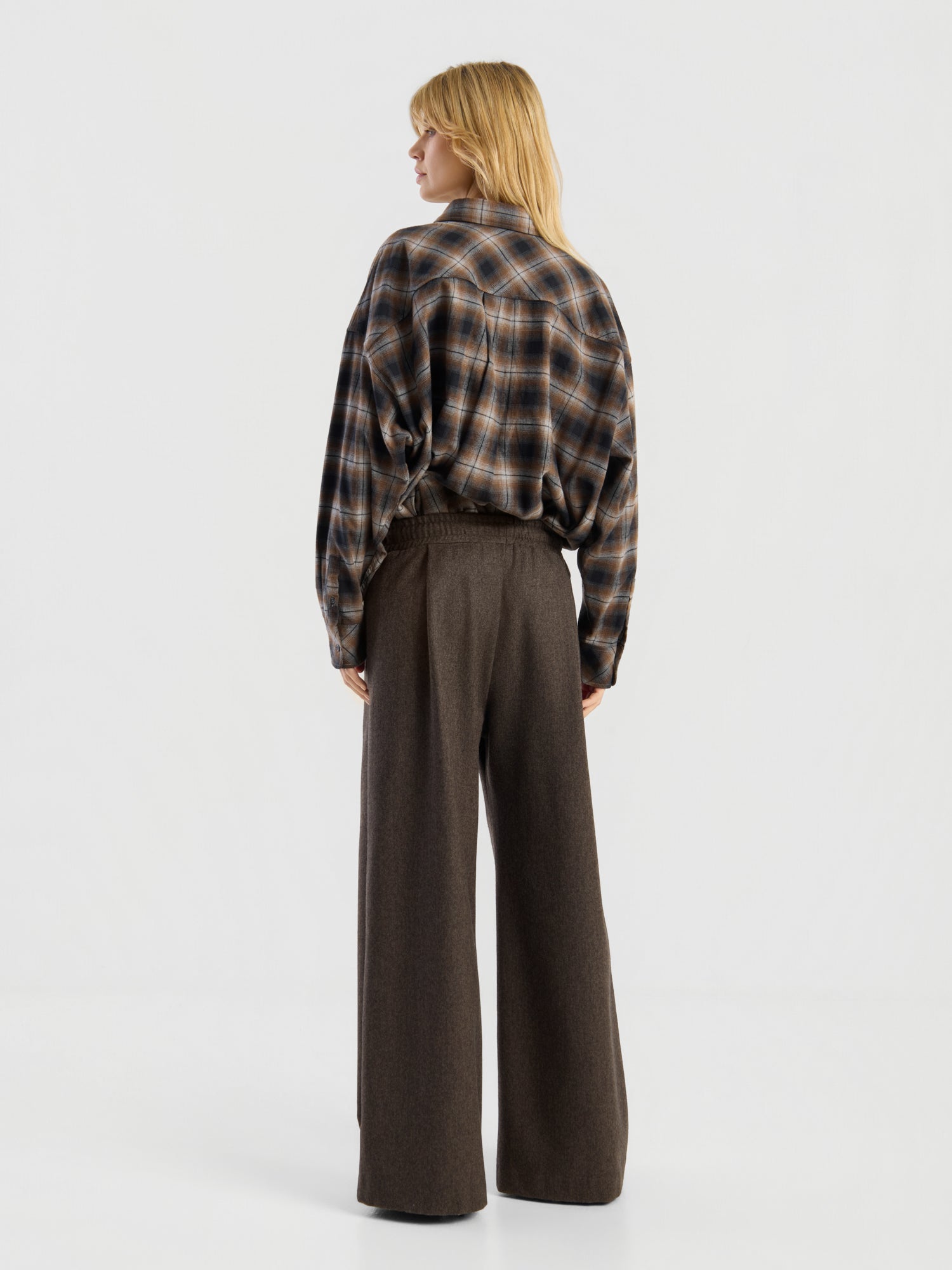 Vor einem schlichten weißen Hintergrund steht eine Person mit blonden Haaren mit dem Rücken zur Kamera. Sie trägt die „Blouse Boxy Long“ in braunem Karomuster, die in eine braune Hose mit hoher Taille und weitem Bein gesteckt ist.