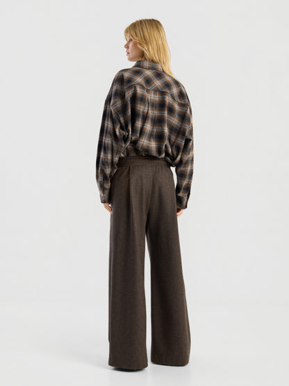 Vor einem schlichten weißen Hintergrund steht eine Person mit blonden Haaren mit dem Rücken zur Kamera. Sie trägt die „Blouse Boxy Long“ in braunem Karomuster, die in eine braune Hose mit hoher Taille und weitem Bein gesteckt ist.