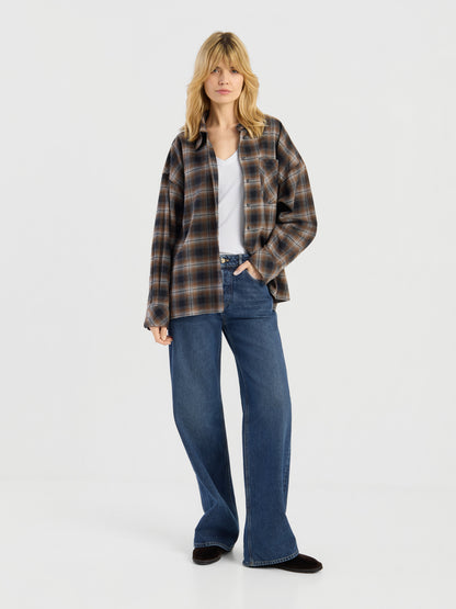 Eine Person mit blonden Haaren trägt die Bluse „Boxy Long“ über einem weißen Oberteil und einer blauen Jeans mit weitem Bein und steht vor einem schlichten weißen Hintergrund.