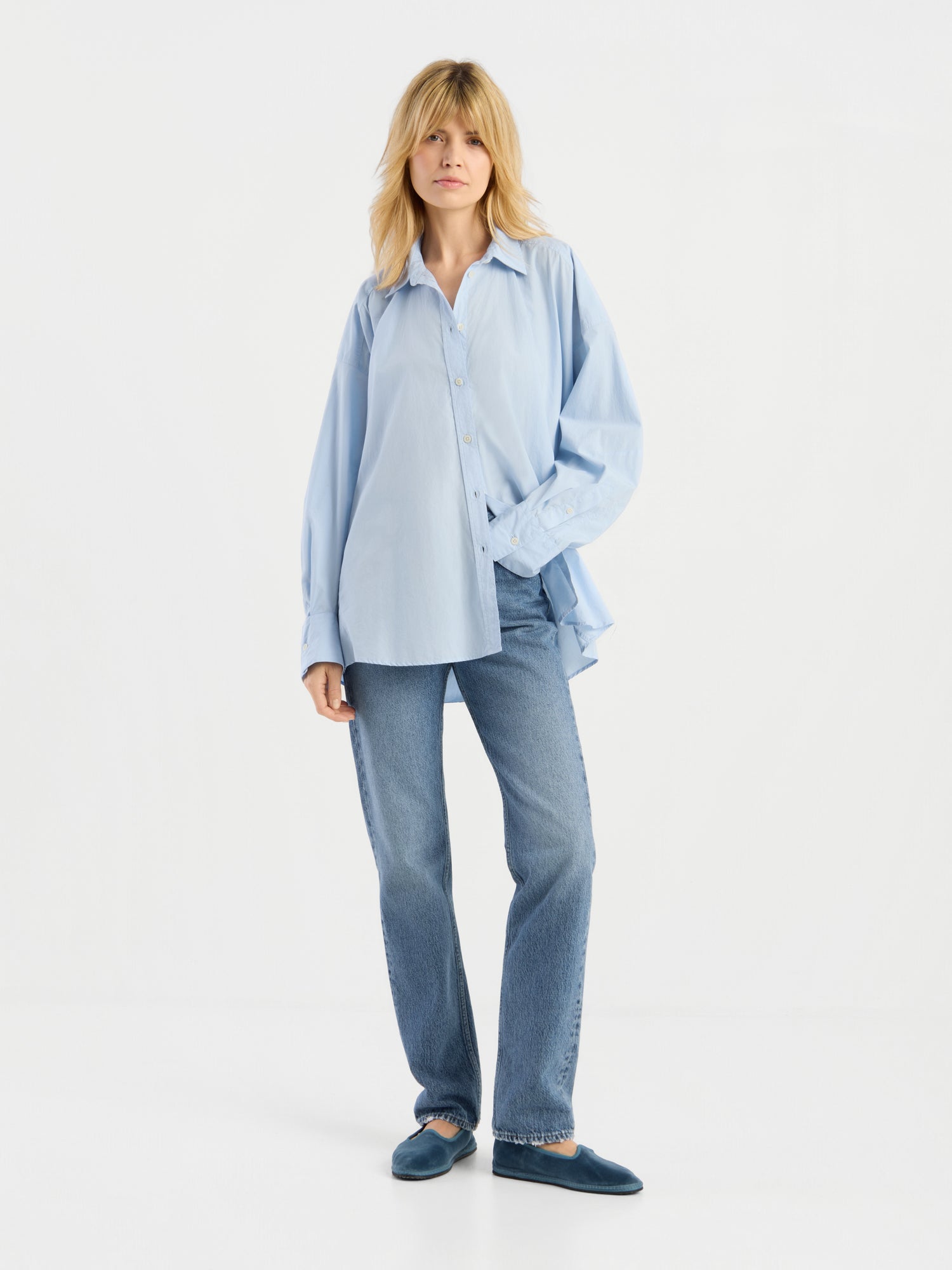 Eine Person mit blonden Haaren trägt ein locker sitzendes, hellblaues Hemd mit Knöpfen, eine „SIENNA Jeans Light Blue – Straight Leg Damenjeans“ sowie blaue Hausschuhe und steht vor einem schlichten weißen Hintergrund.