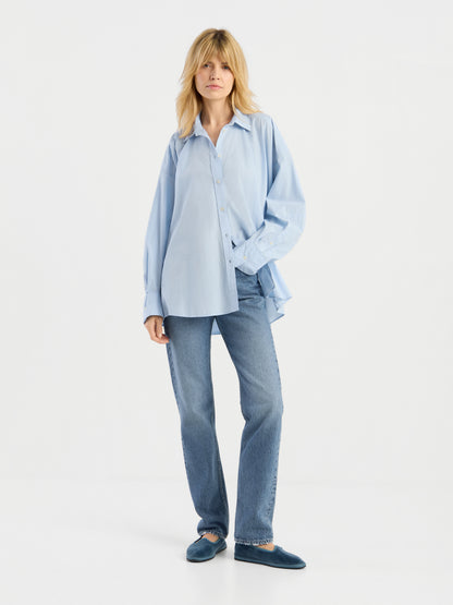 Eine Person mit blonden Haaren trägt ein locker sitzendes, hellblaues Hemd mit Knöpfen, eine „SIENNA Jeans Light Blue – Straight Leg Damenjeans“ sowie blaue Hausschuhe und steht vor einem schlichten weißen Hintergrund.