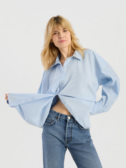 Eine Person mit blonden Haaren trägt die Bluse „Hannah Lightblue“ und eine blaue Jeans und lächelt, während sie den Saum ihrer Bluse vor einem schlichten, hellen Hintergrund hochhält.
