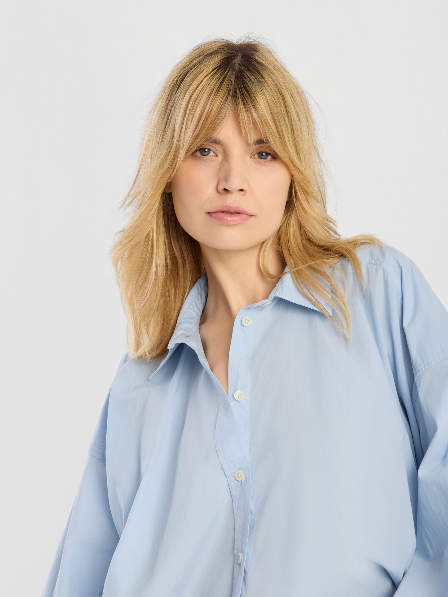 Eine Frau mit blonden Haaren und Pony trägt die Bluse „Hannah Lightblue“, steht vor einem schlichten hellen Hintergrund und blickt mit neutralem Gesichtsausdruck leicht zur Seite.