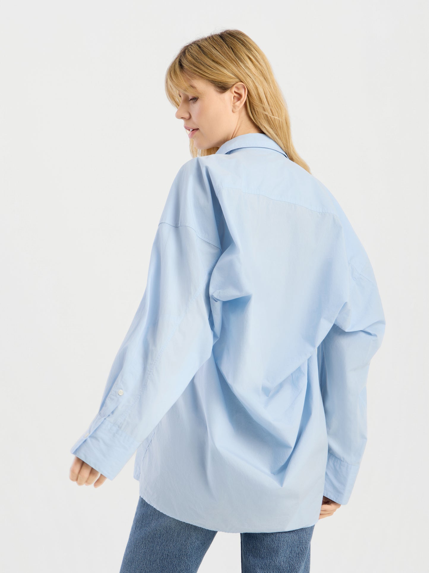 Eine Frau mit blonden Haaren präsentiert die Bluse „Hannah Lightblue“ in Kombination mit einer blauen Jeans; sie steht seitlich und blickt nach unten, vor einem schlichten weißen Hintergrund.