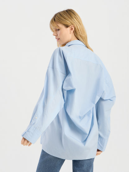 Eine Frau mit blonden Haaren präsentiert die Bluse „Hannah Lightblue“ in Kombination mit einer blauen Jeans; sie steht seitlich und blickt nach unten, vor einem schlichten weißen Hintergrund.