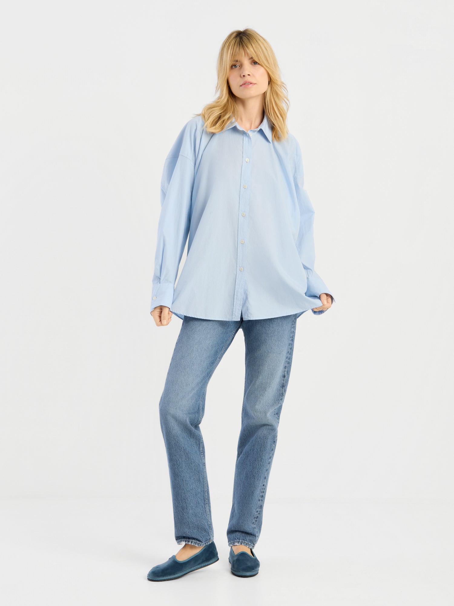 Eine Frau mit blonden Haaren trägt die Bluse „Hannah Lightblue“ locker geschnitten, dazu eine gerade geschnittene blaue Jeans und blaue Slipper. Sie steht vor einem schlichten weißen Hintergrund und hält die Seiten ihrer Bluse fest.