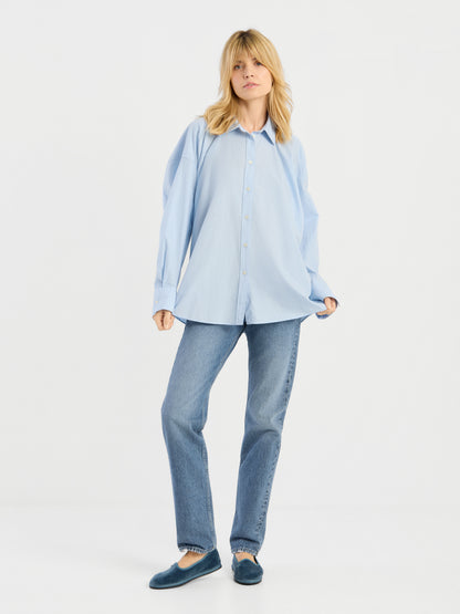 Eine Frau mit blonden Haaren trägt die Bluse „Hannah Lightblue“ locker geschnitten, dazu eine gerade geschnittene blaue Jeans und blaue Slipper. Sie steht vor einem schlichten weißen Hintergrund und hält die Seiten ihrer Bluse fest.