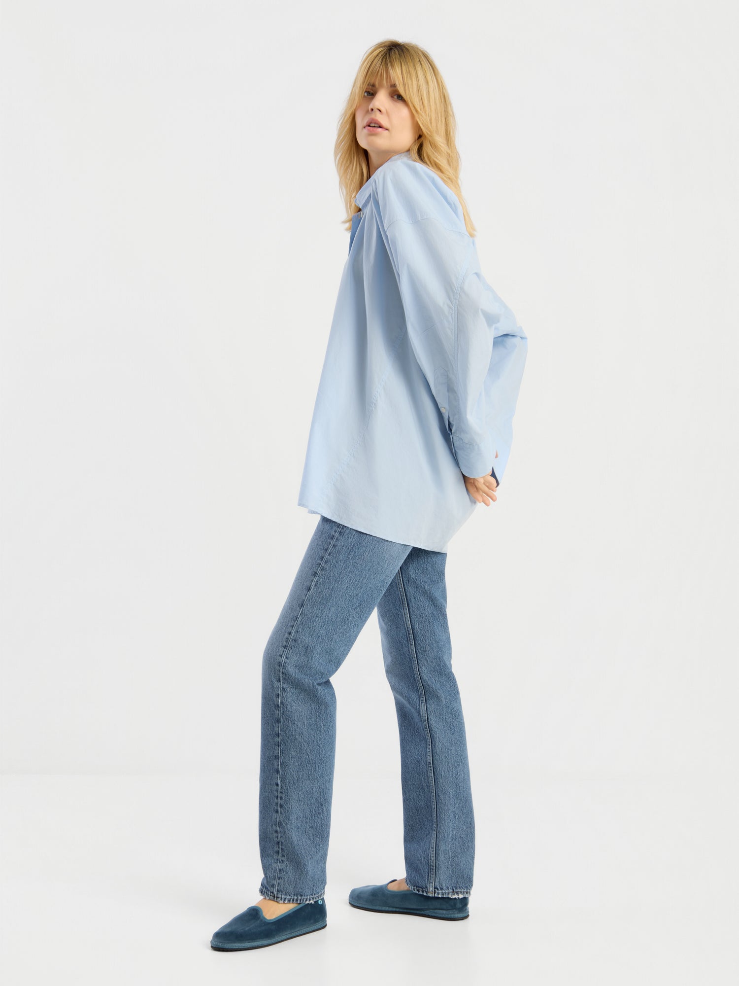 Eine Person mit langen blonden Haaren trägt die Bluse „Hannah Lightblue“, eine blaue Jeans und blaue Hausschuhe und steht seitlich mit hinter dem Rücken verschränkten Armen vor einem weißen Hintergrund.