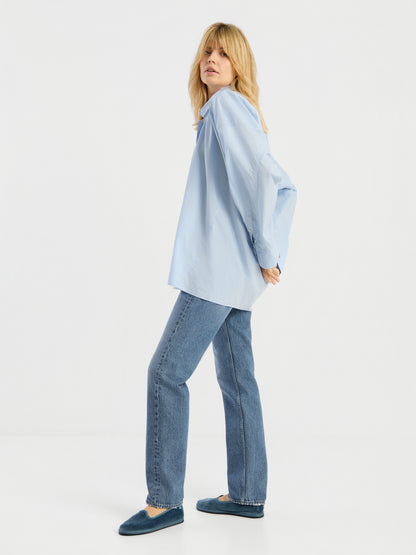 Eine Person mit langen blonden Haaren trägt die Bluse „Hannah Lightblue“, eine blaue Jeans und blaue Hausschuhe und steht seitlich mit hinter dem Rücken verschränkten Armen vor einem weißen Hintergrund.