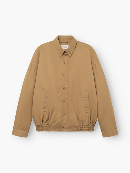 Beigefarbene Bomber-Shorts mit Knopfleiste, Kragen, elastischem Saum und zwei Vordertaschen, abgebildet vor weißem Hintergrund.