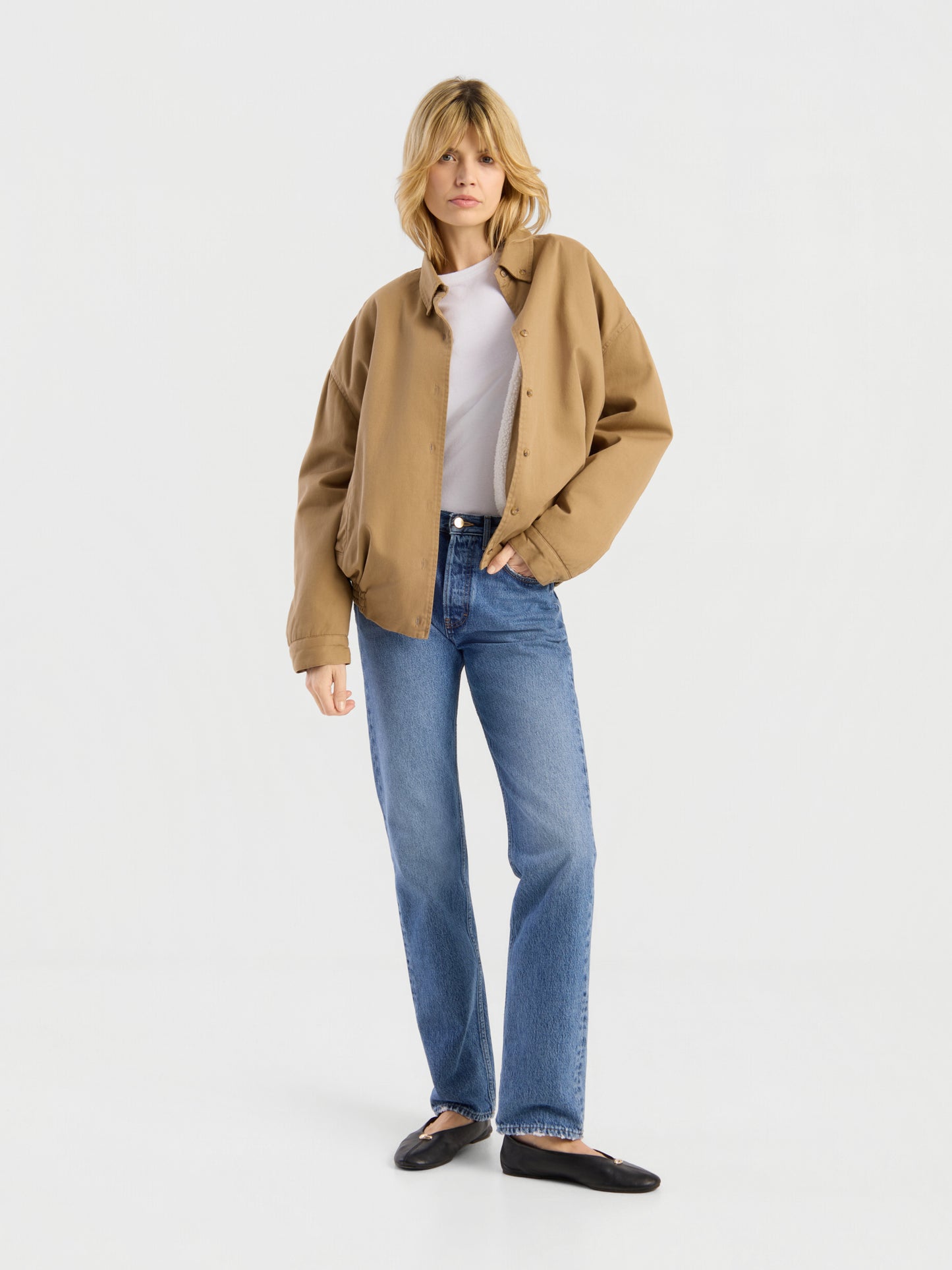 Eine Person mit blonden Haaren trägt die beigefarbene „Button Down Bomber Short“-Jacke über einem weißen Hemd, blauen Jeans und schwarzen Schuhen und steht vor einem schlichten weißen Hintergrund.