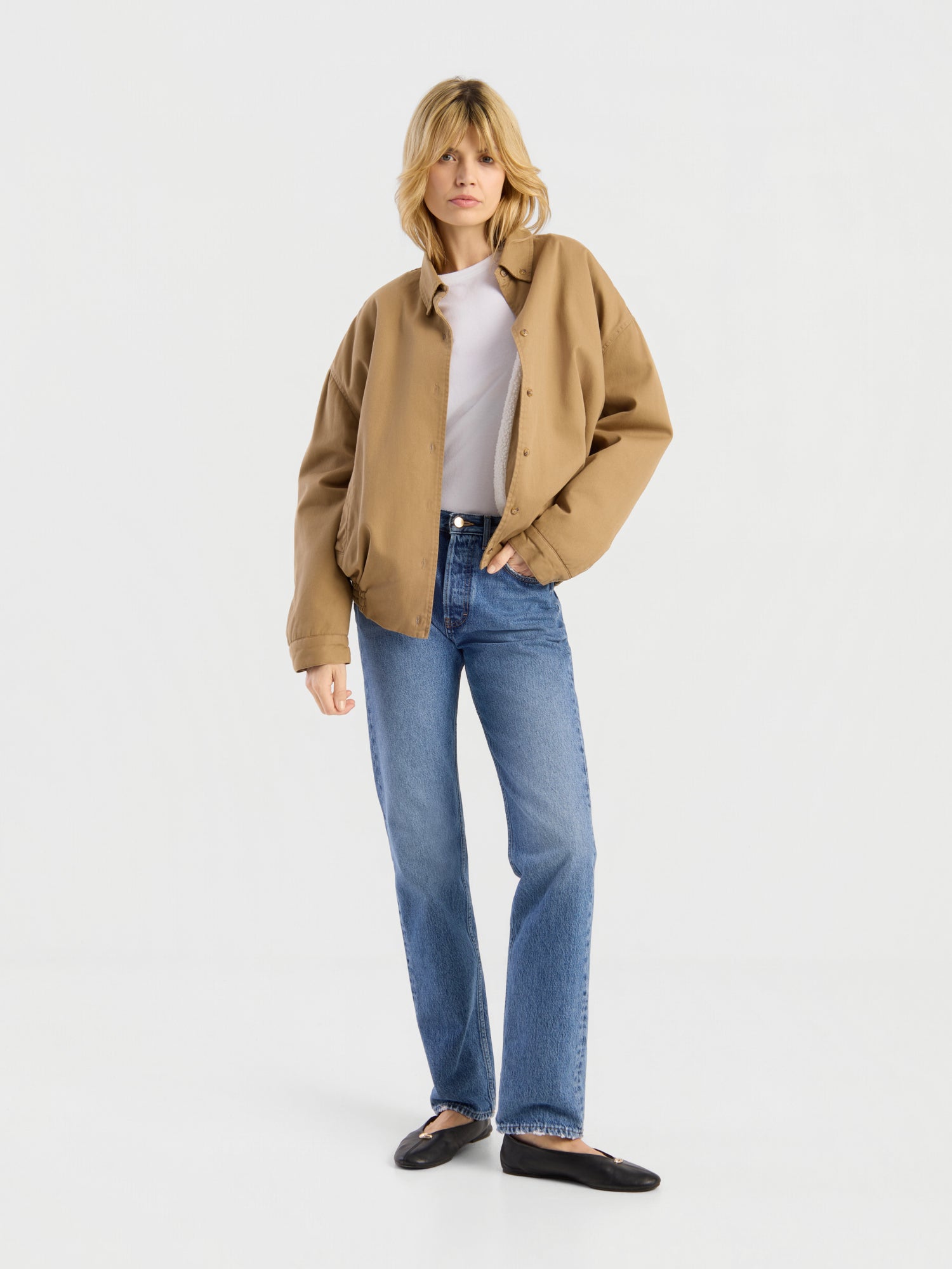 Eine Person mit blonden Haaren trägt die beigefarbene „Button Down Bomber Short“-Jacke über einem weißen Hemd, blauen Jeans und schwarzen Schuhen und steht vor einem schlichten weißen Hintergrund.