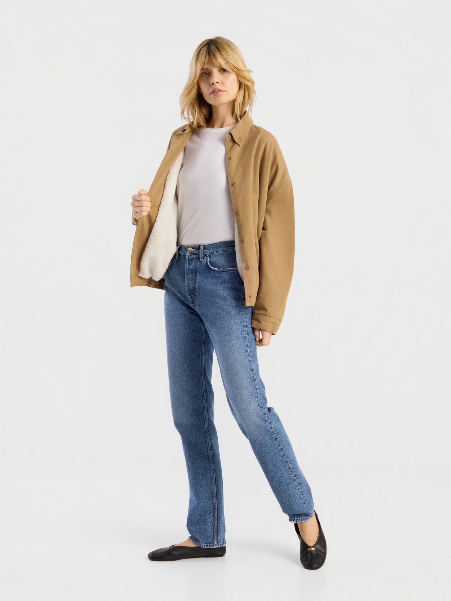 Eine Frau steht vor einem schlichten weißen Hintergrund. Sie trägt eine hellbraune Jacke, ein weißes Hemd und schwarze Schuhe und präsentiert die „SIENNA Jeans Light Blue – Straight Leg“-Damenjeans.
