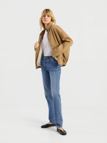 Eine Person steht vor einem weißen Hintergrund und trägt die beigefarbene „Button Down Bomber Short“-Jacke über einem weißen Hemd, dazu blaue Jeans und schwarze Ballerinas; sie blickt nach vorne und hält die Jacke mit einer Hand offen.